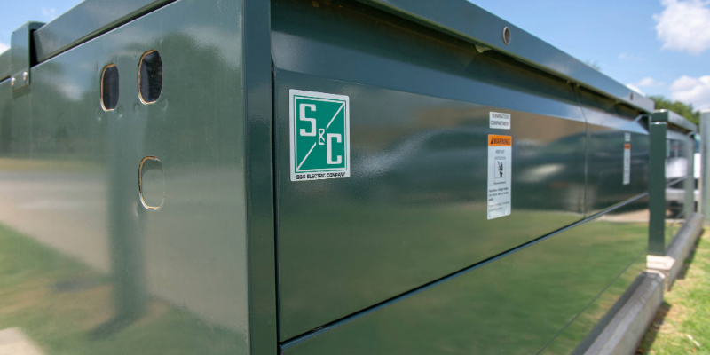 Closeup of one corner of S&C&rsquo;s Vista Underground Distribution Switchgear. The green S&C logo is in the foreground. In the background is a blue sky with puffy white clouds.
