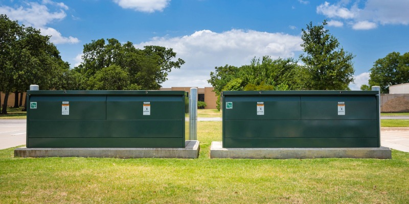 Two green underground distribution switchgear boxes on concrete pads. In the foreground is green grass. In the background are trees and a blue sky