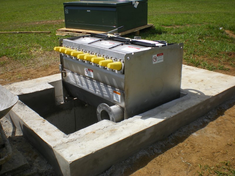 A Vista® Underground Distribution Switchgear unit is being installed on a concrete pad in the foreground. The sealed metal interior of the unit is exposed, and the green enclosure that will cover it sits on a wood pallet on the grass in the background. The unit has nine protruding bushings covered by yellow caps. Its base rests on a dug-out part of the concrete pad, and beneath it is a further dug-out space of unknown depth. 