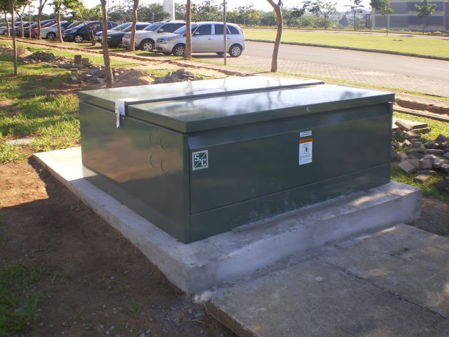 The fully installed Vista switchgear unit, complete with its dark green S&C-branded enclosure, sits on its concrete pad in the foreground. Evidence of the installation work is present, including a now-empty wood pallet in the foreground, freshly dug dirt around the concrete pad, and scattered materials on the grass. In the background are a row of trees, a row of parked vehicles, and a road.